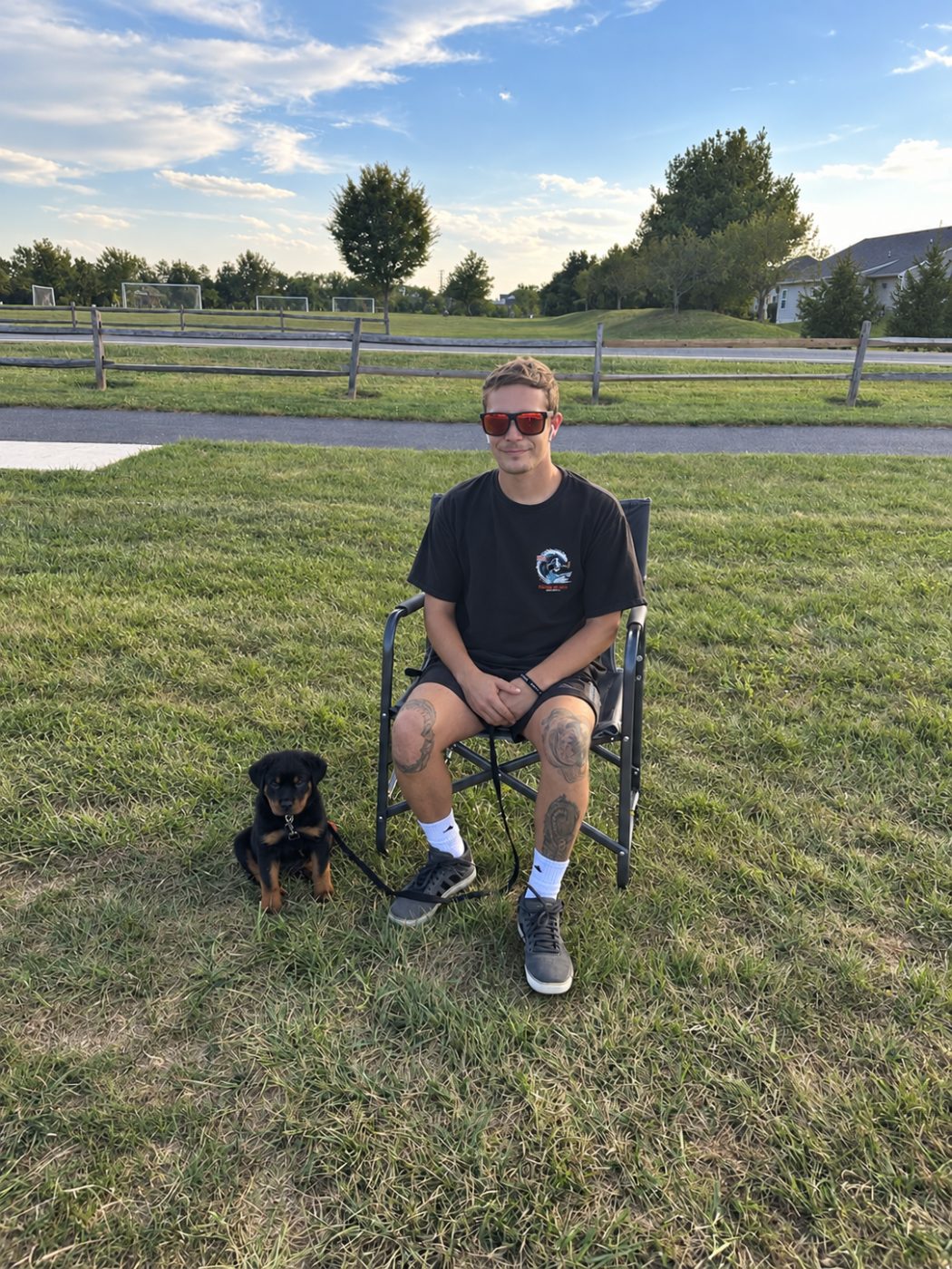 Alex sitting in a camp chair on a grassy field beside Kaiju, his Rottweiler puppy