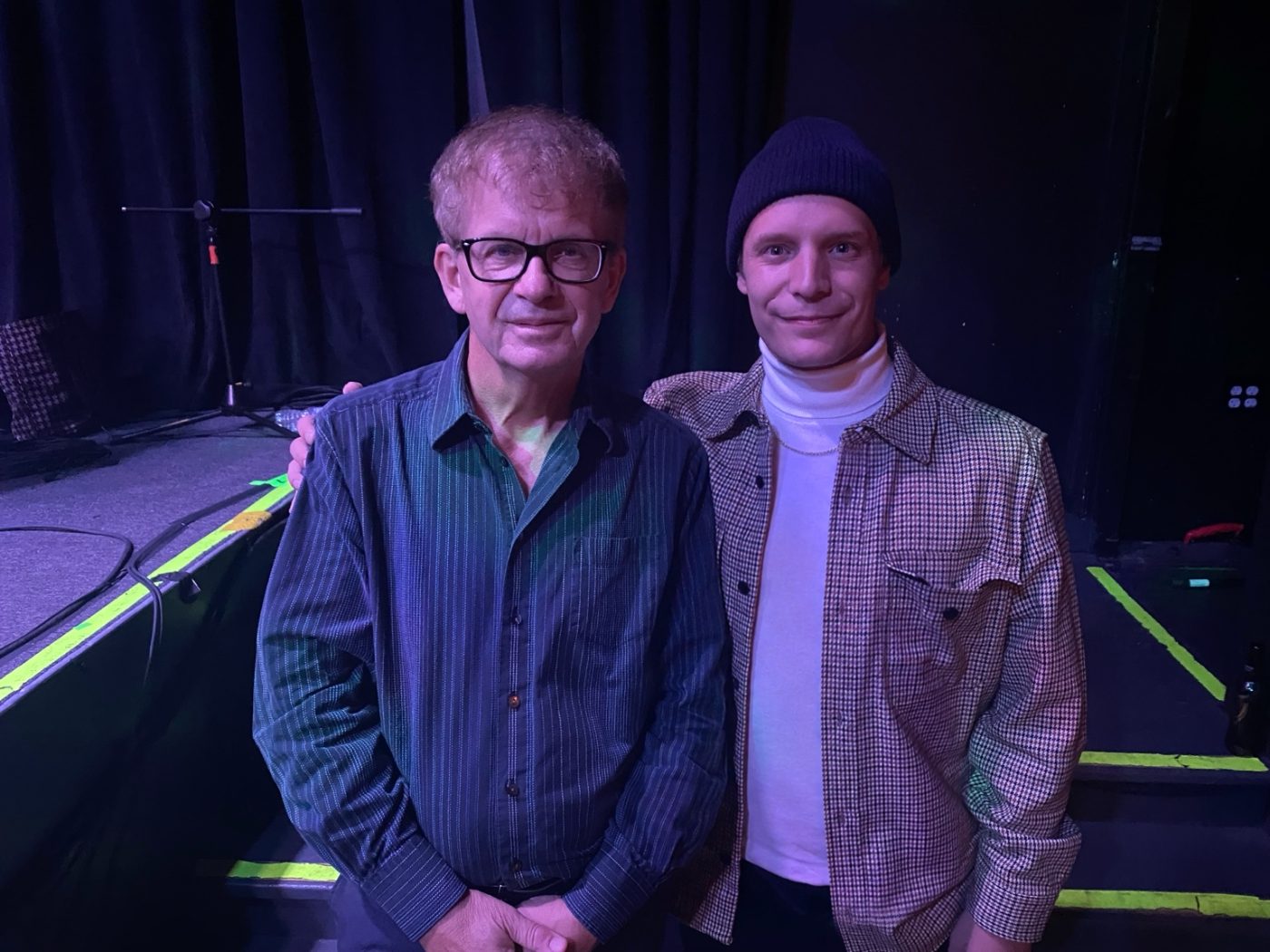 Alex standing beside a musician at the edge of a stage after a show
