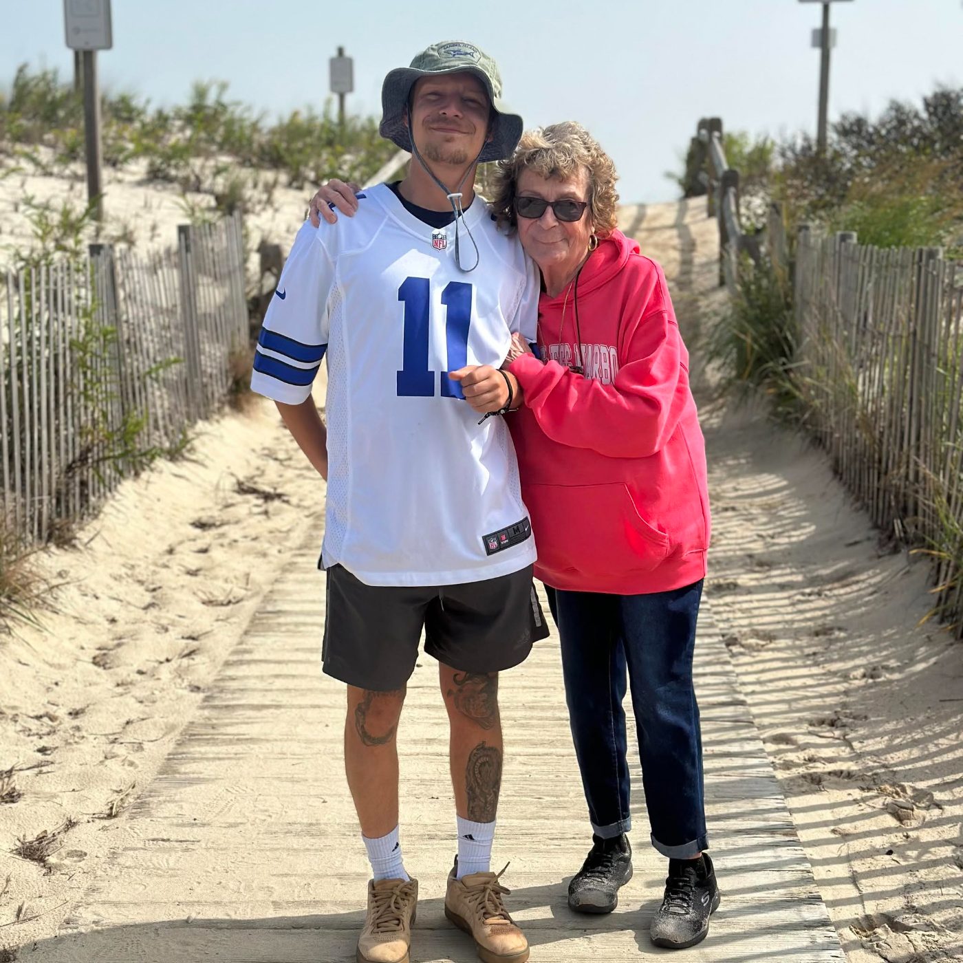Alex in a Dallas Cowboys jersey standing with his grandmother on a boardwalk through beach dunes